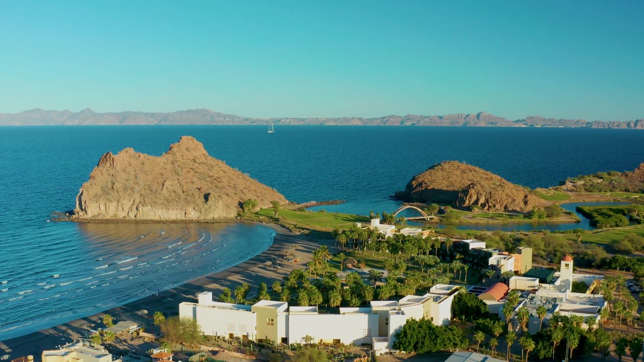 Opening aerial frame over Loreto, Baja California Sur shows a tan rock outcrop beside a curved beach, blue ocean, white coastal buildings, palms, distant mountains, and no visible people.