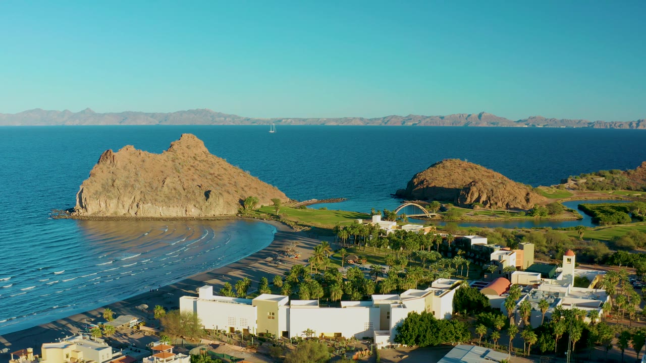 Loreto, Baja California Sur coastline appears from above with the large rock on the left, a smaller rocky rise to the right, low white buildings below, blue water behind, and no visible people.