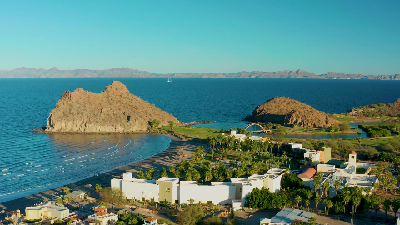 Aerial shoreline frame with blue bay, rocky outcrop, beach, palms, and white buildings.