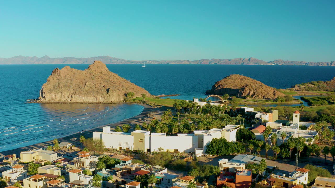 Opening aerial frame over Loreto, Baja California Sur shows red-roofed homes, a large white building, palm trees, curved beach, blue ocean, tan rock outcrops, distant mountains, and no visible people.