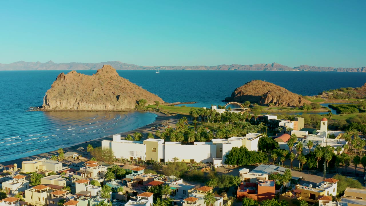 At 0:02, the aerial frame shows Loreto rooftops and palms below, a large white structure near the beach, rippled blue water, two tan rocky outcrops, distant mountains, and no visible people.