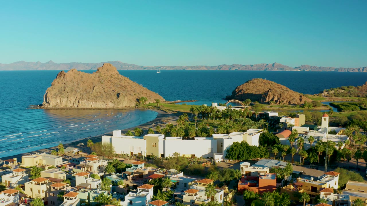 Aerial coastline view with small wave lines, white buildings, palms, and rocky headlands.