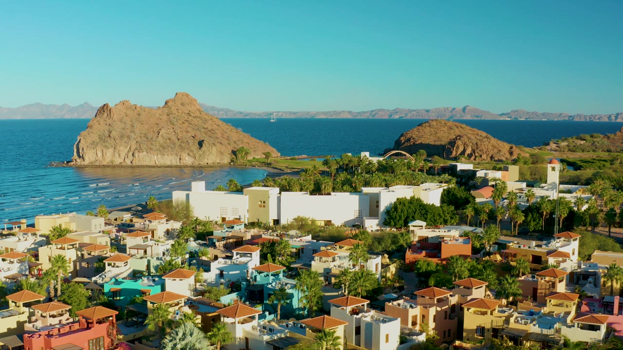 Loreto aerial frame with sunlit houses and palms in the foreground, white buildings near the shore, small waves along the beach, rocky formations offshore, distant mountains, and no visible people.