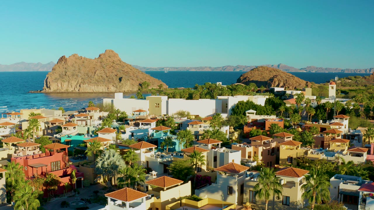 Opening frame of Loreto, Baja California Sur, showing red tile roofs, palms, blue ocean, a rocky outcrop offshore, distant mountains, and no visible people.