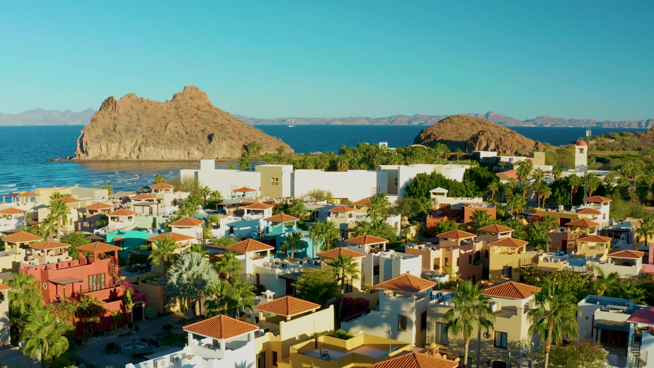 Aerial composition over Loreto homes shows clustered palms, bright white buildings, blue water, a tan rocky outcrop, and no visible people.