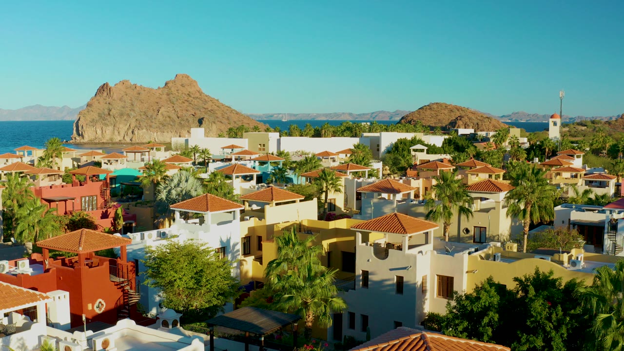 Wide aerial view over Loreto, Baja California Sur rooftops with palms, blue water, a rocky island, and no visible people.