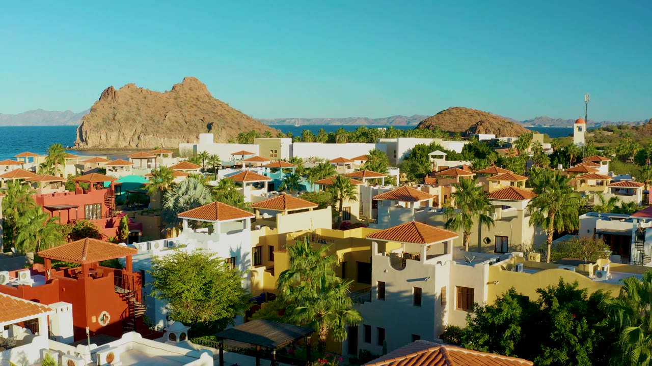 Aerial view across low town buildings with red roofs, palms, sea, and rocky island.