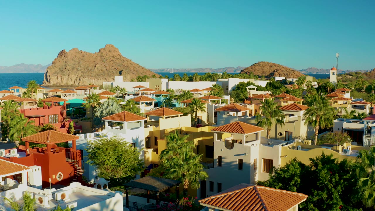 Loreto coastal neighborhood seen from above with warm sun on rooftops, palms in the midground, blue water, rocky island, and no visible people.