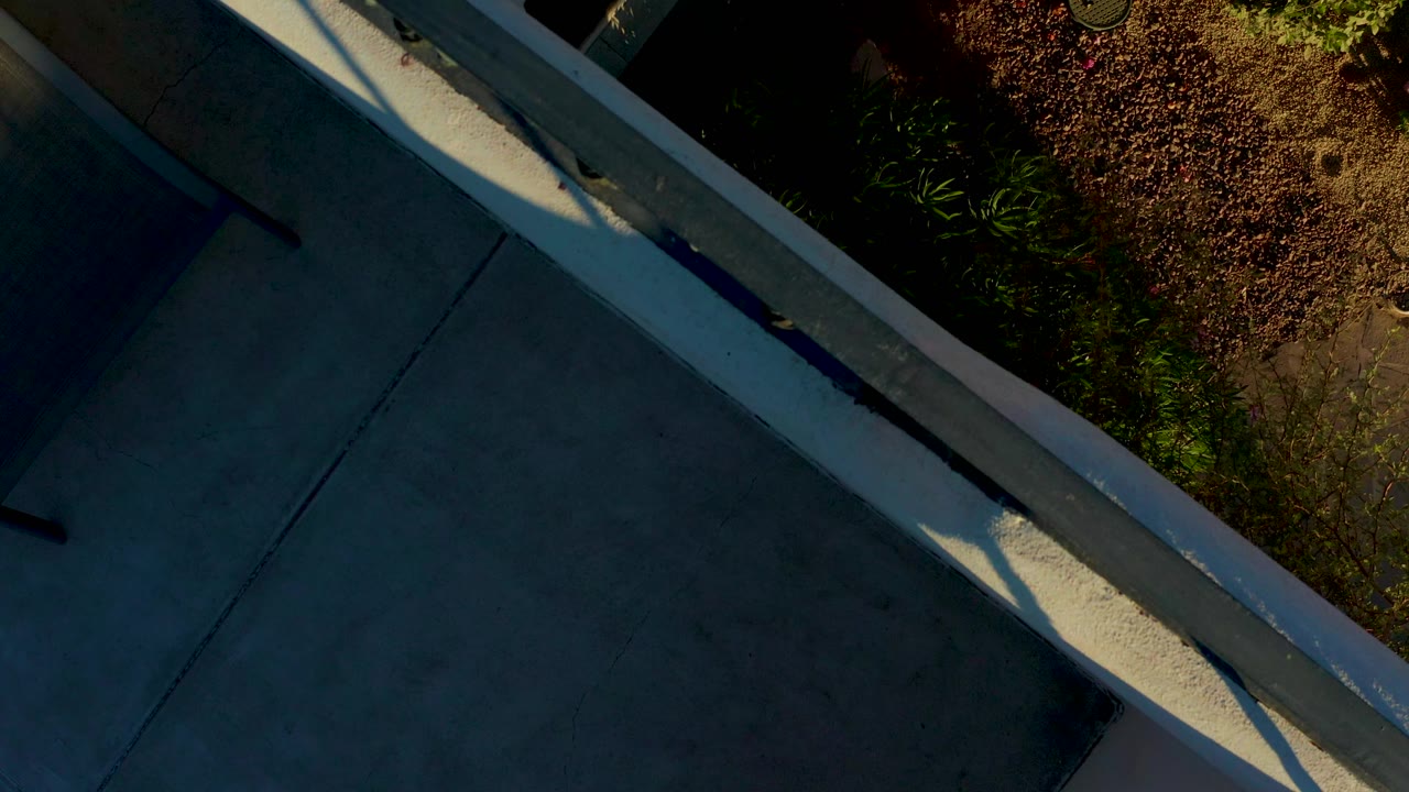 Overhead view of a concrete ledge and railing in Loreto, Baja California Sur, with dark paved surface, planting at the edge, strong shadows, and no visible people.