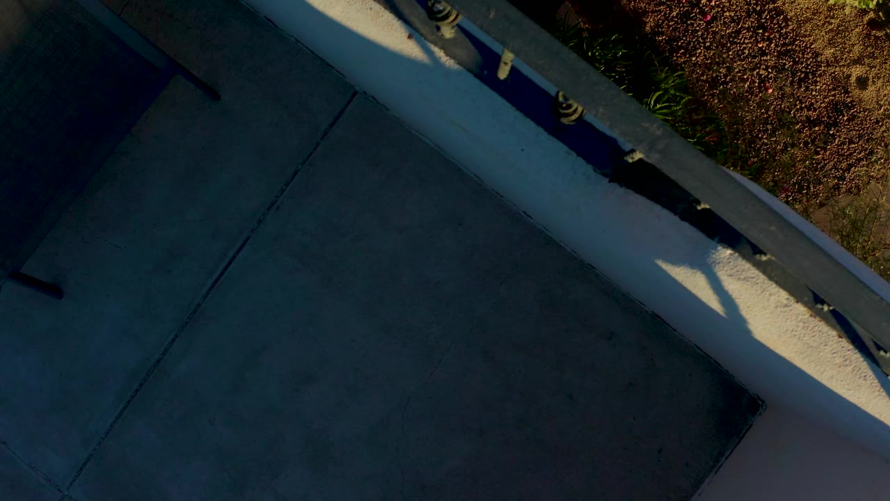 The elevated view remains tight on the concrete ledge and railing in Loreto, with landscaped plants along the upper edge, hard sunlight, and no visible people.
