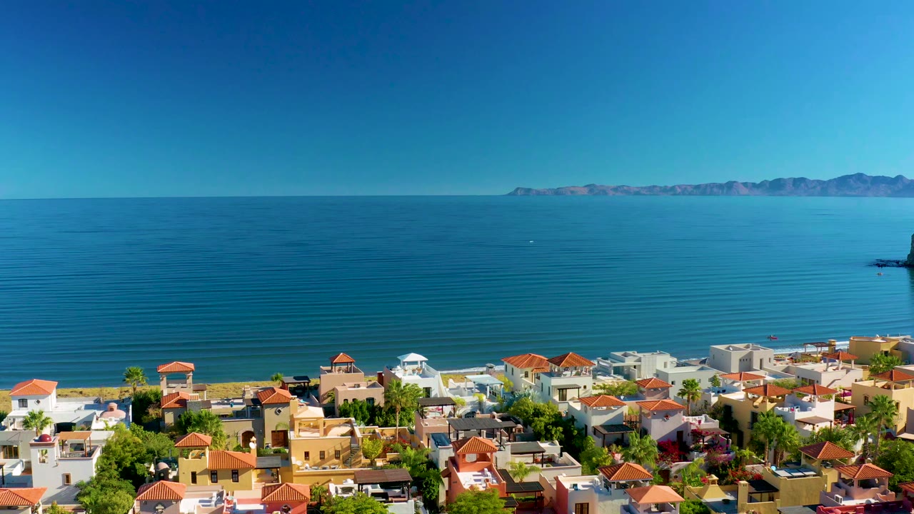 Opening aerial frame over Loreto, Baja California Sur, showing red-tile roofs, white and tan buildings, palms, sandy shoreline, calm blue ocean, distant mountains, and no visible people.