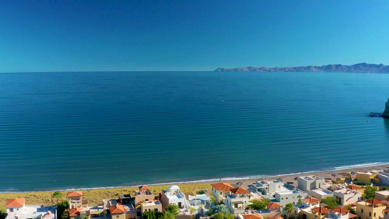 Aerial frame over Loreto, Baja California Sur, with blue ocean filling most of the view, a sandy beach and coastal homes along the bottom, tiny people visible near the shore, and distant mountains on the horizon.