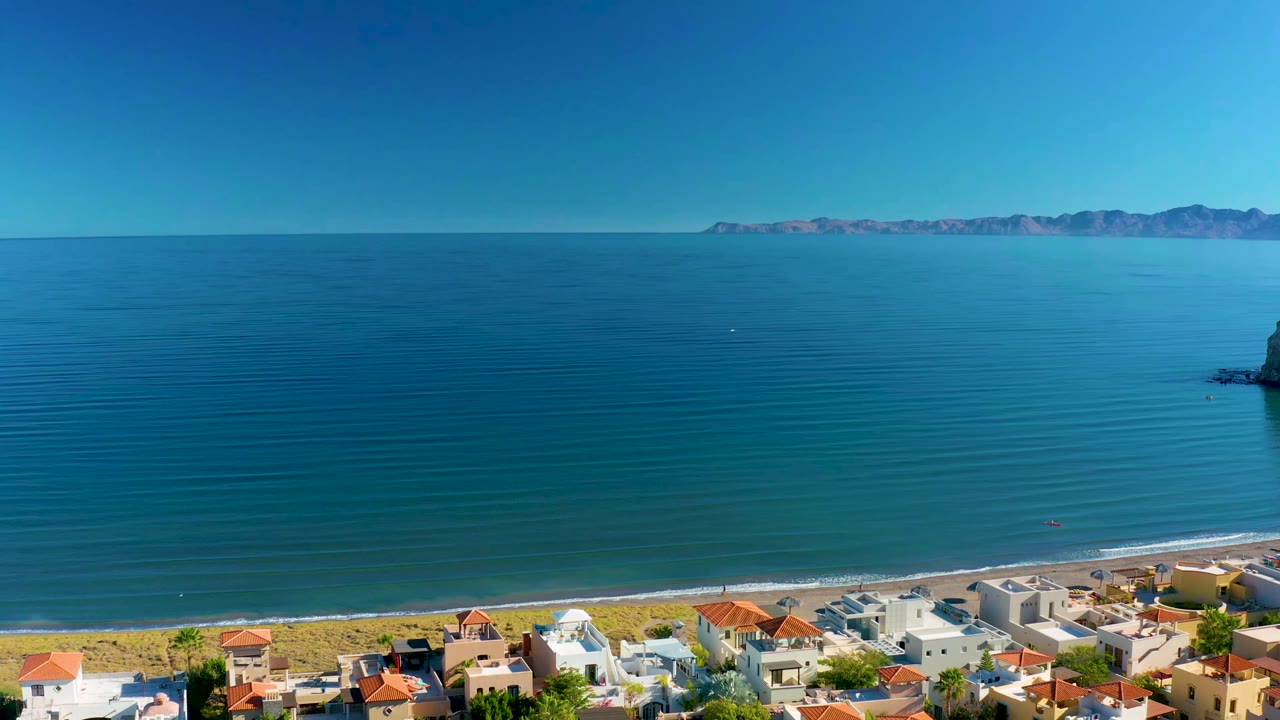 Wide drone view of calm water, beach, and red-roofed buildings.