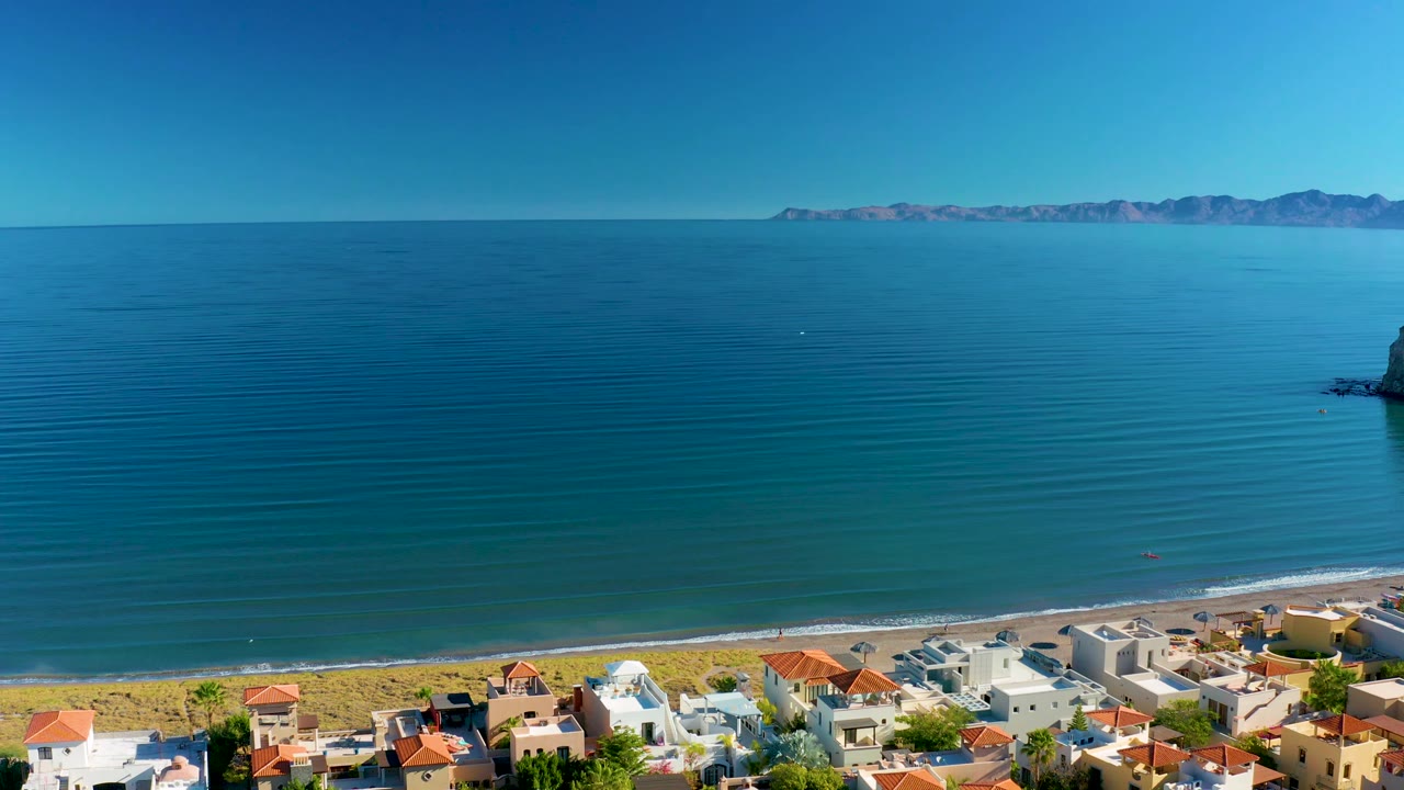 High aerial view over Loreto with deep blue coastal water, a thin surf line, beach and low homes at the bottom of frame, tiny beachgoers visible, and a mountain range faintly lining the horizon.