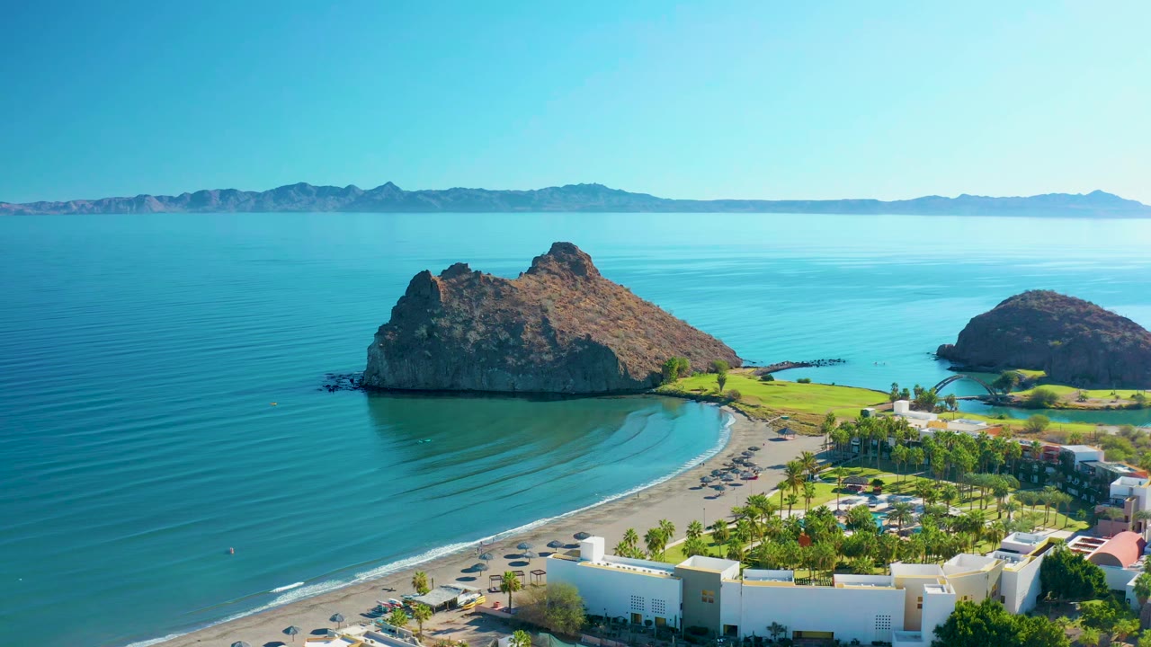 Opening aerial frame over Loreto, Baja California Sur, Mexico, showing a rocky island, curved sandy beach, shore buildings, distant mountains, calm blue water, and no visible people.