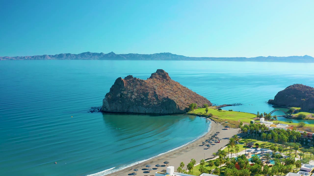 Aerial view in Loreto, Baja California Sur, with the rocky island centered beside the beach, parallel water ripples offshore, a small parking area near palms, and no visible people.