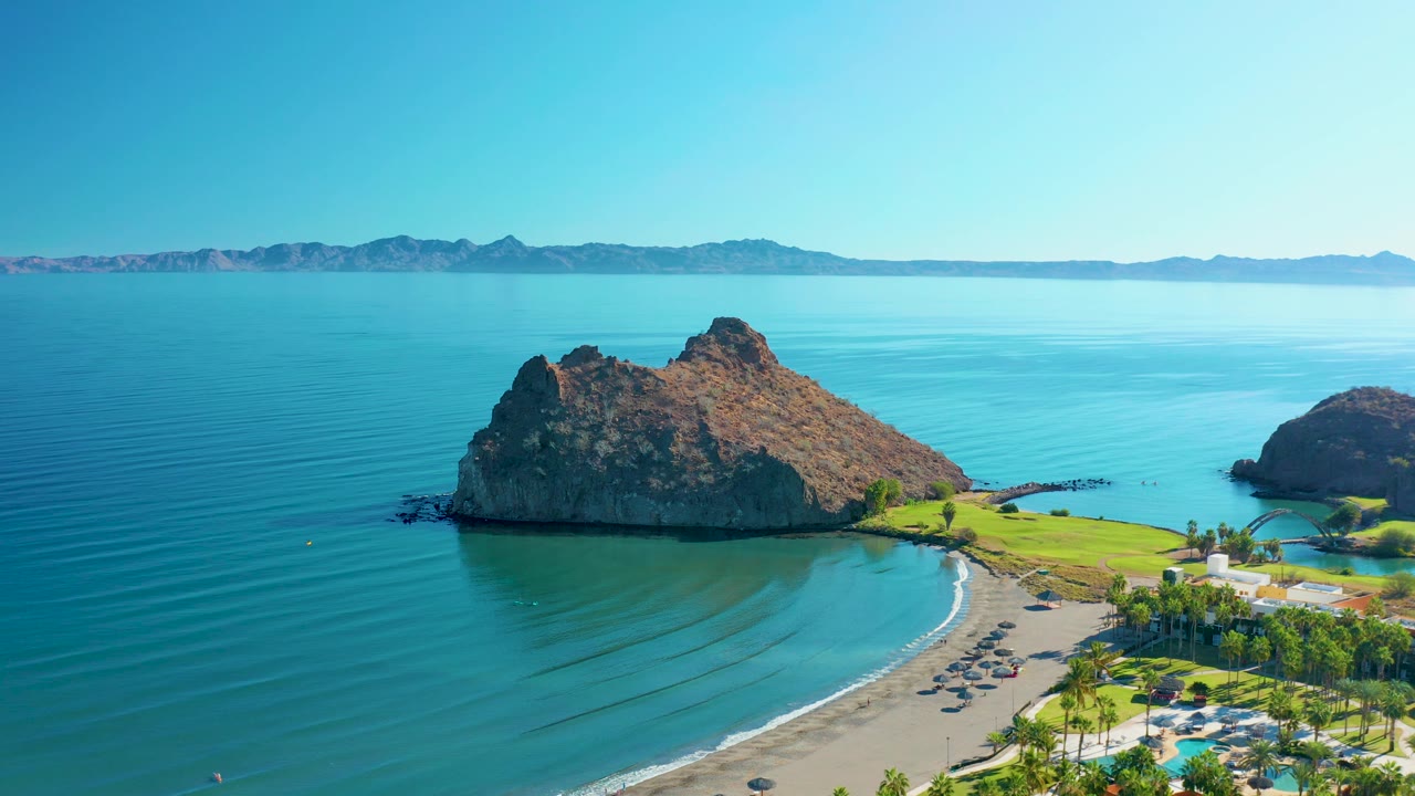Aerial frame over Loreto, Baja California Sur, with the rocky outcrop closer in frame, curved surf line on the beach, parked cars near palm trees, distant mountains, and no visible people.