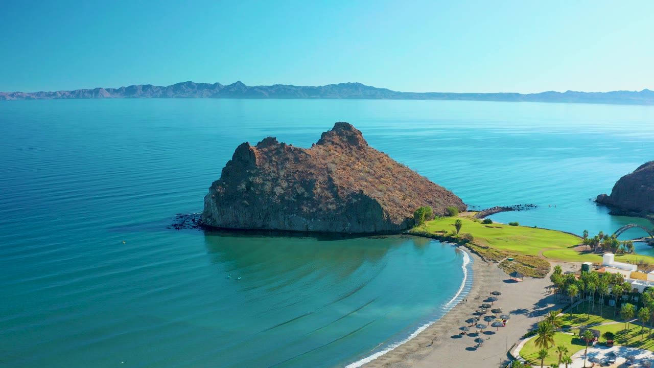 Aerial frame from Loreto, Baja California Sur showing a rocky island, curved sandy beach, green shoreline, distant mountains, and no visible people.
