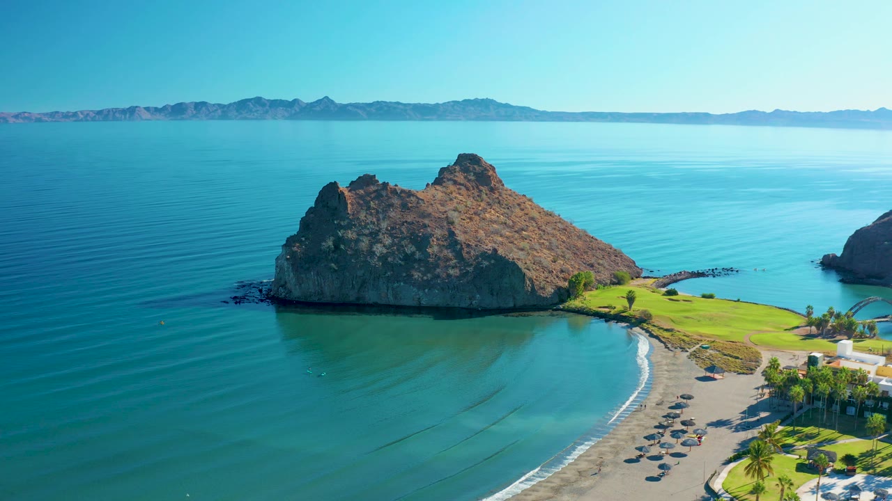 Loreto aerial frame showing the rocky island slightly right of center, smooth blue ocean, the curved beach below, and no visible people.