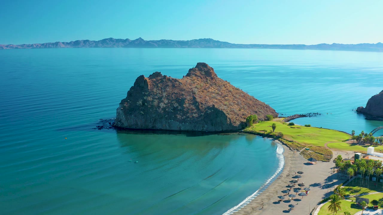 Aerial view over Loreto coastline with the rocky island above the beach curve, narrow white surf lines, distant mountains, and no visible people.
