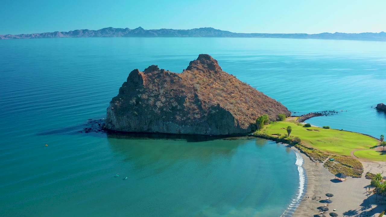 Aerial view in Loreto, Baja California Sur, Mexico, showing a rocky outcrop beside turquoise water, a sandy beach, a strip of green grass, distant mountains, and no visible people.