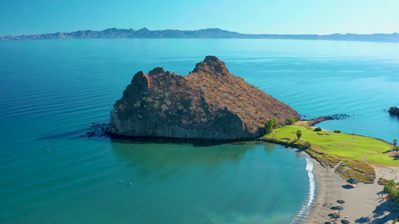A high aerial frame in Loreto holds the rocky outcrop near center, with blue-green water below, a sandy beach curving at right, green grass beyond it, and no visible people.