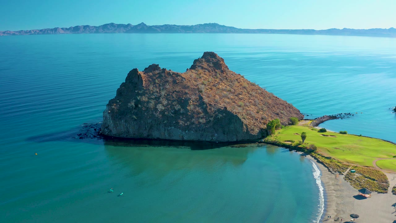 Aerial shoreline view with rocky outcrop, calm sea, beach, and grass.