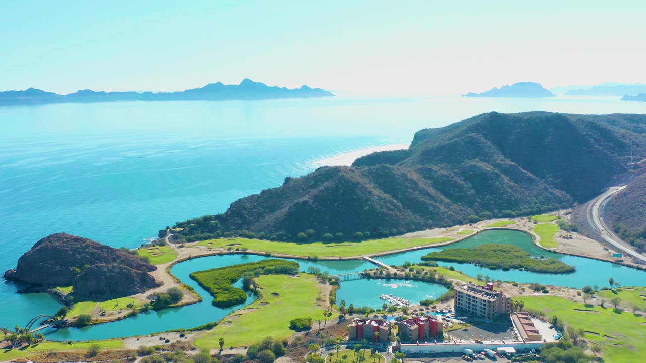 Wide aerial of coastal hills, blue water, green inlets, road, buildings, and islands on the horizon.