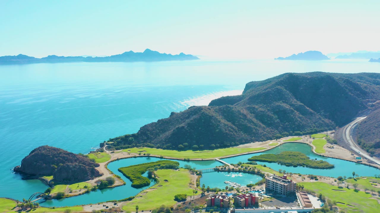 High aerial view over Loreto, Baja California Sur, showing a turquoise lagoon, green grounds, coastal hills, a curving road, distant landforms, and no visible people.