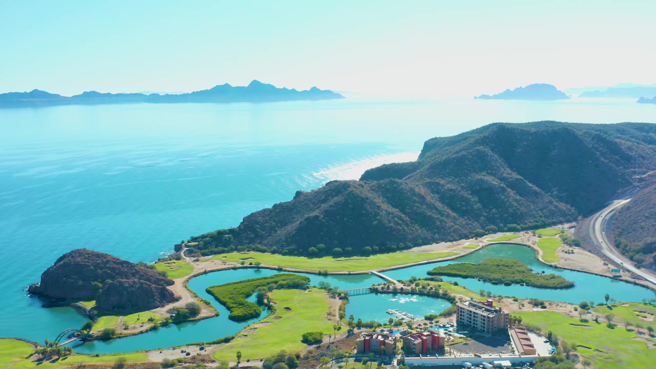 Loreto aerial view showing the marina area and buildings near the bottom edge, lagoon water and grass in the center, coastal hills and road to the right, and no visible people.