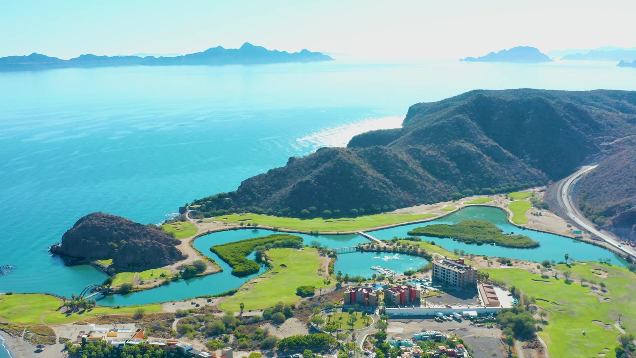 High aerial frame over Loreto, Baja California Sur with turquoise ocean, distant islands, dark coastal hills, green golf fairways, winding lagoon water, roads, buildings, cars, and no visible people.