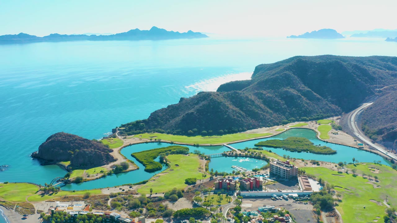 Drone view of coastal golf greens, lagoon channels, hills, road, ocean, and buildings.
