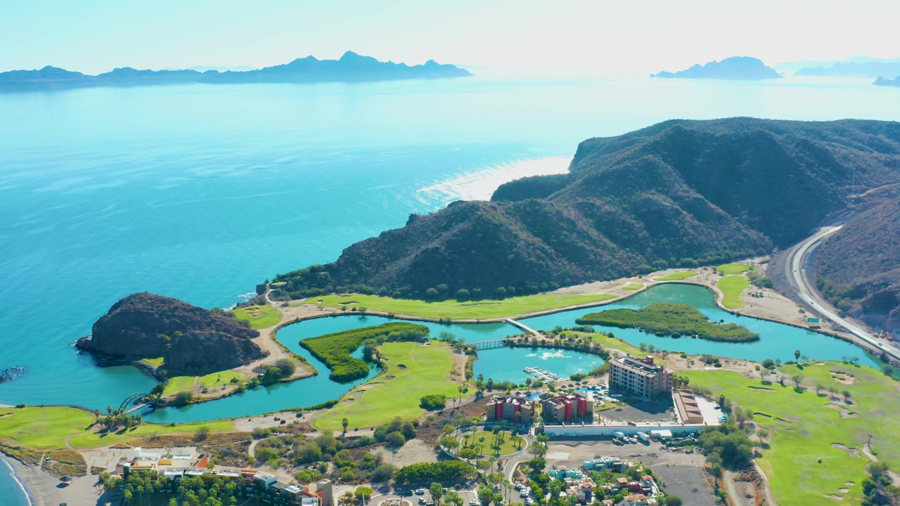 Loreto coastal aerial with blue sea and islands across the horizon, hills above a curving road, green fairways, lagoon water, marina-like docks, buildings, cars, and no visible people.