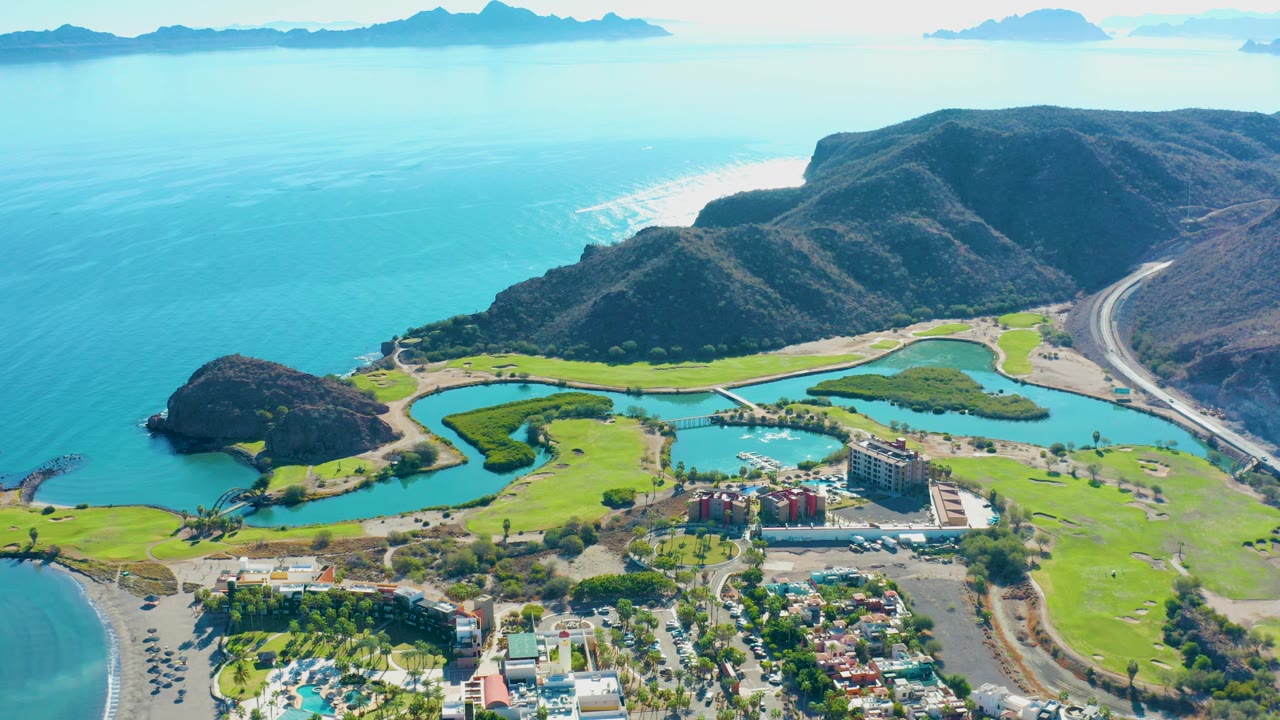 Aerial coastal golf course with ocean, mountains, road, and buildings.