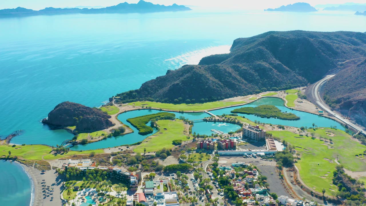 High aerial frame of Loreto with the golf course and lagoon-like water features centered below, blue ocean to the left, brown mountain ridges behind, and no visible people.