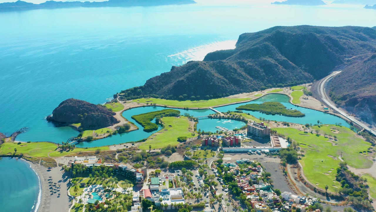Aerial frame over Loreto with a curved beach at lower left, green course land and water features through the center, a road along the mountain side, and no visible people.
