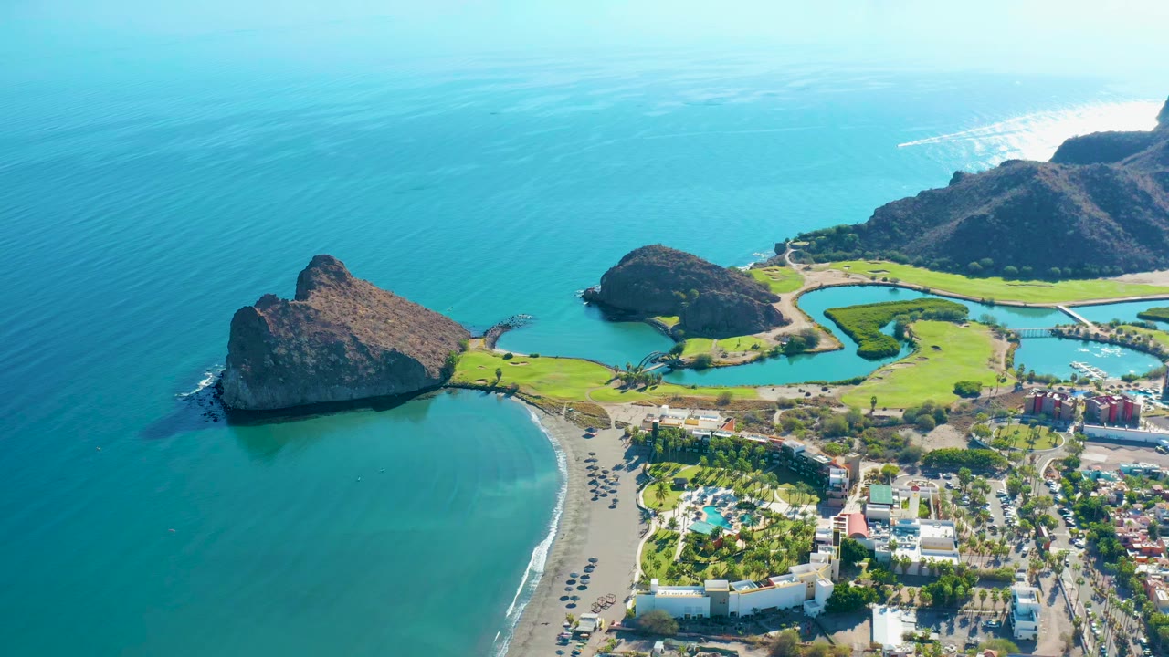 Opening aerial frame over Loreto, Baja California Sur, with turquoise water at left, a curved sandy beach below, rocky headlands, green golf fairways, shoreline buildings, parked cars, and no visible people.