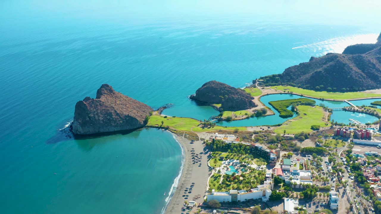 Aerial frame in Loreto, Baja California Sur, with the beach curving around shallow green-blue water, rocky headlands set against the sea, a golf course and buildings inland, and no visible people.