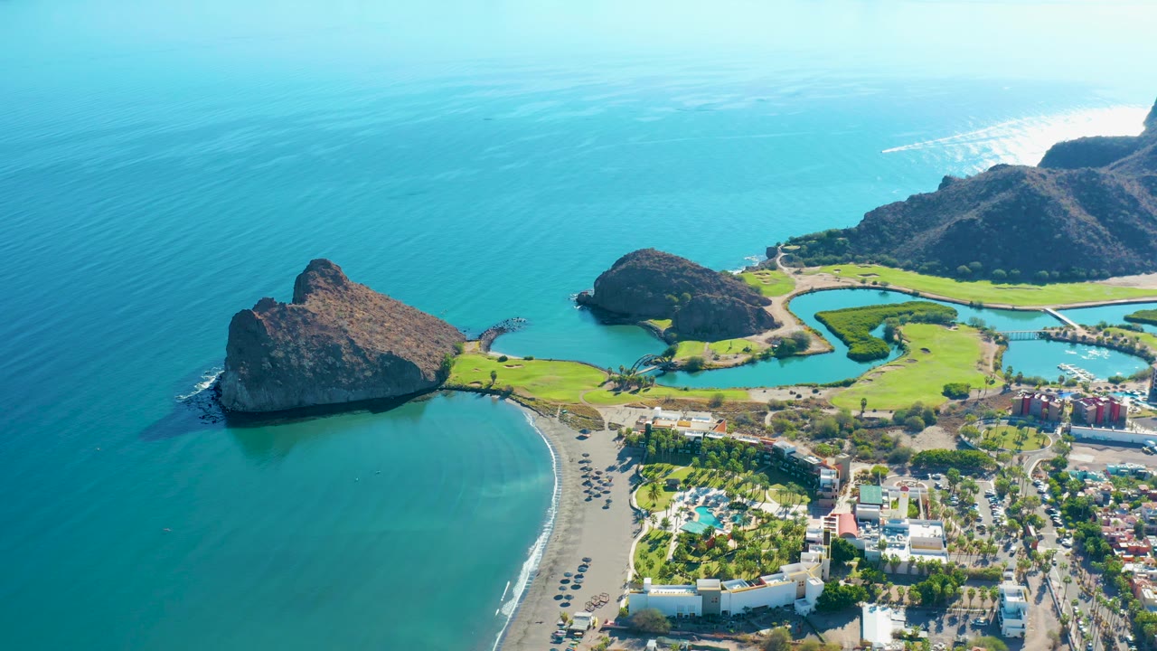 Aerial shoreline scene with turquoise water, rocky outcrop, sand beach, golf course, and distant hills.
