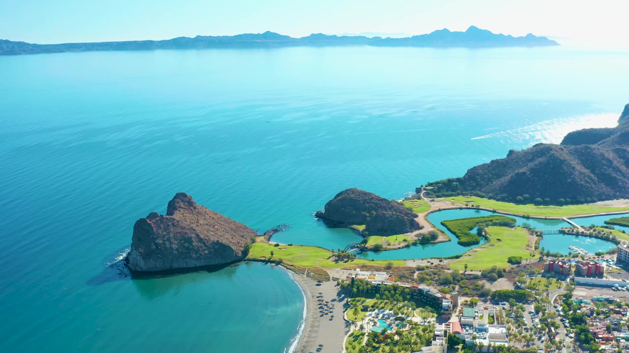 Aerial view in Loreto, Baja California Sur with the rocky point and beach lower in frame, green fields and buildings to the right, hazy mountain horizon, and no visible people.