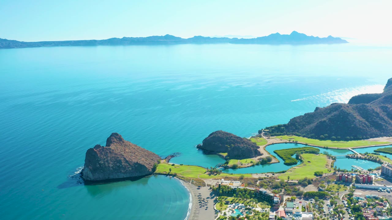 Mid-clip aerial view over Loreto, Baja California Sur with open blue sea across most of the frame, rocky outcrop near the beach, green shoreline areas, buildings, distant mountains, and no visible people.