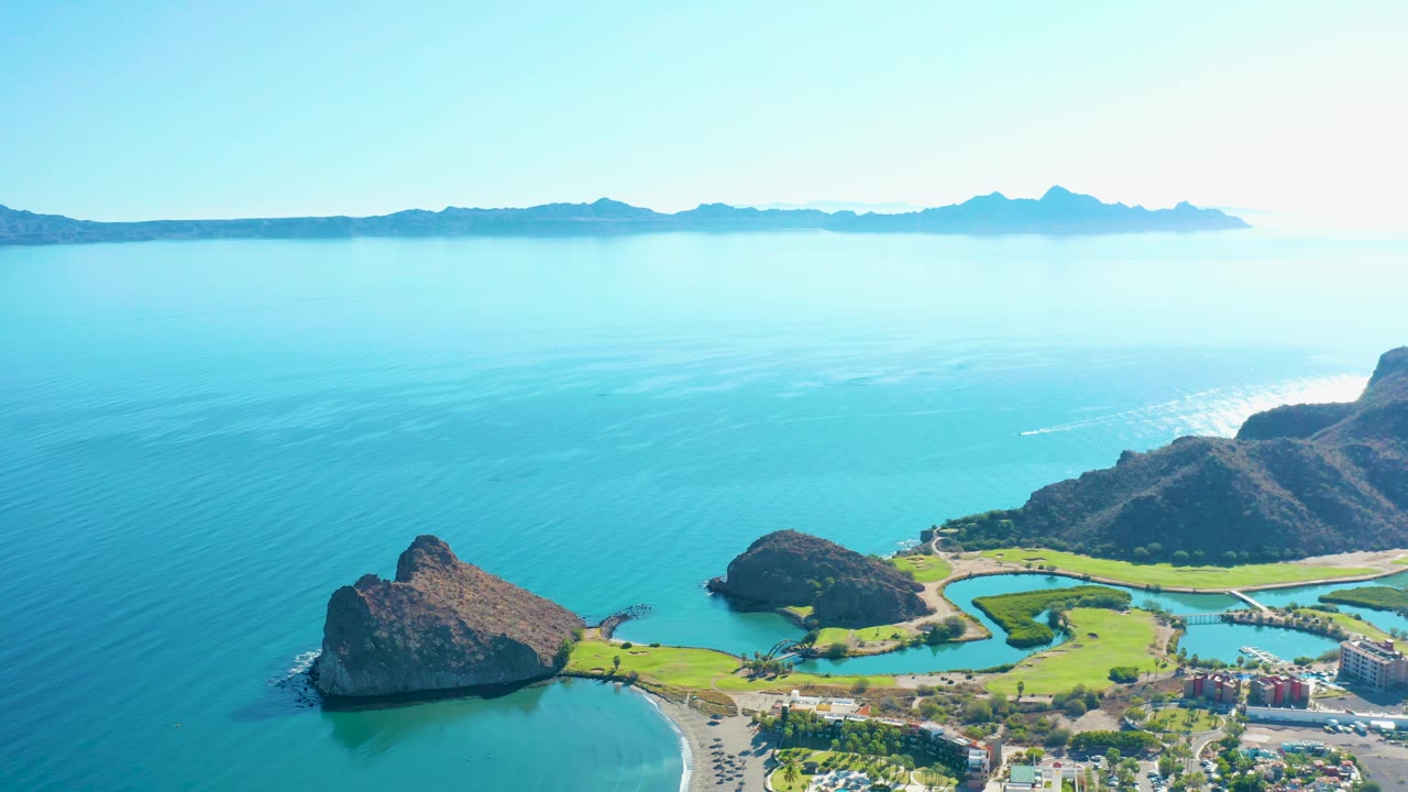 A second aerial frame of Loreto, Baja California Sur keeps the rocky points and developed shoreline low in frame with calm blue water and distant mountains behind, no visible people.