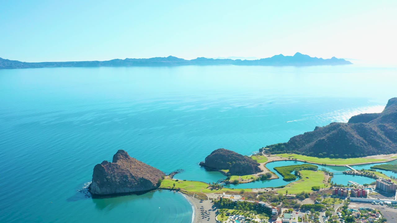 Aerial view over Loreto, Baja California Sur with turquoise water filling the frame, green shore development at the lower right, rocky cliffs, a beach, and no visible people.