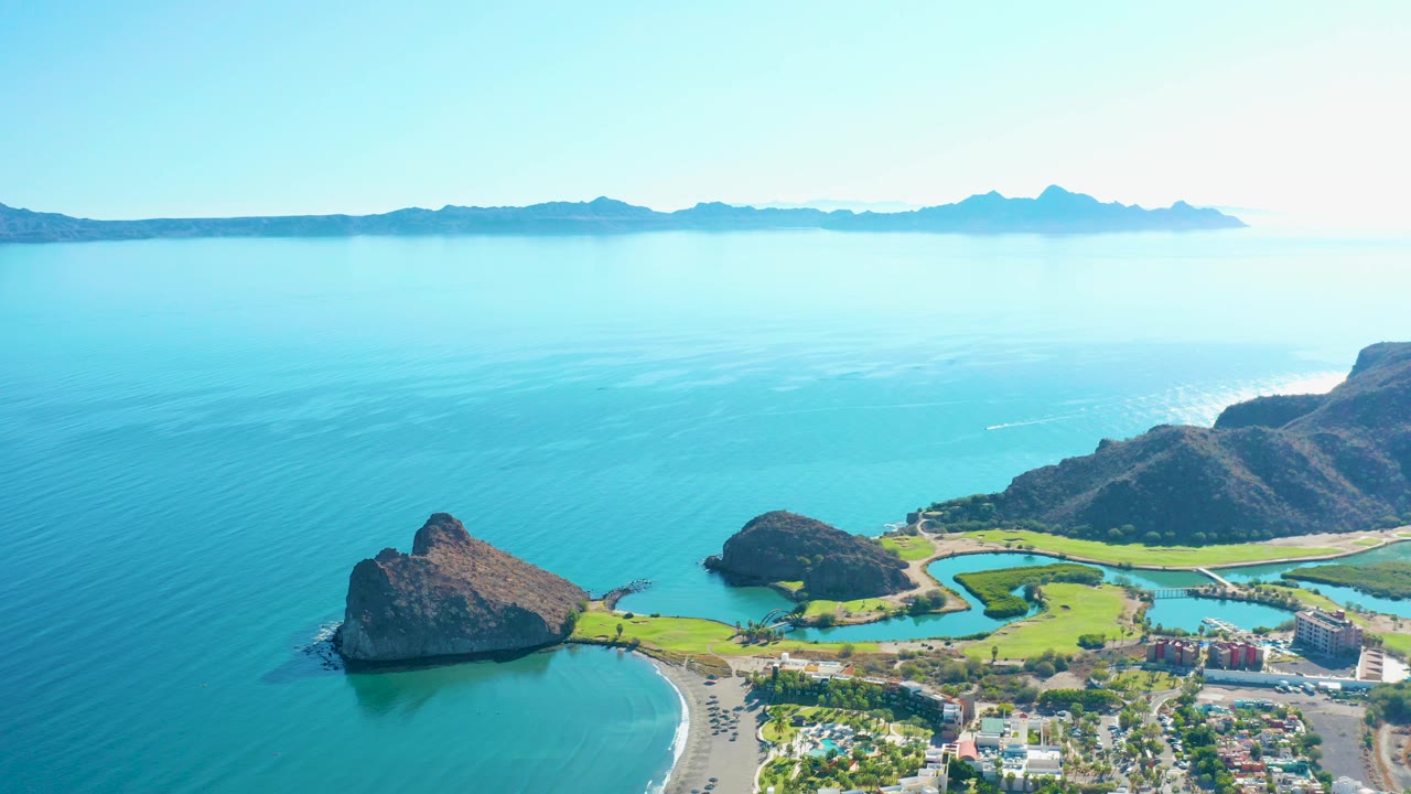 Opening frame over Loreto, Baja California Sur shows turquoise water around a sandy beach, rocky coastal hills, green shoreline, town buildings, distant mountains, and no visible people.