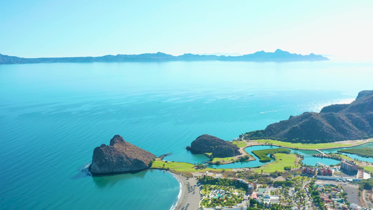 At 0:02, the Loreto shoreline remains bright and open, with calm turquoise ocean, beach sand, green coastal land, rocky hills to the right, town buildings below, and no visible people.