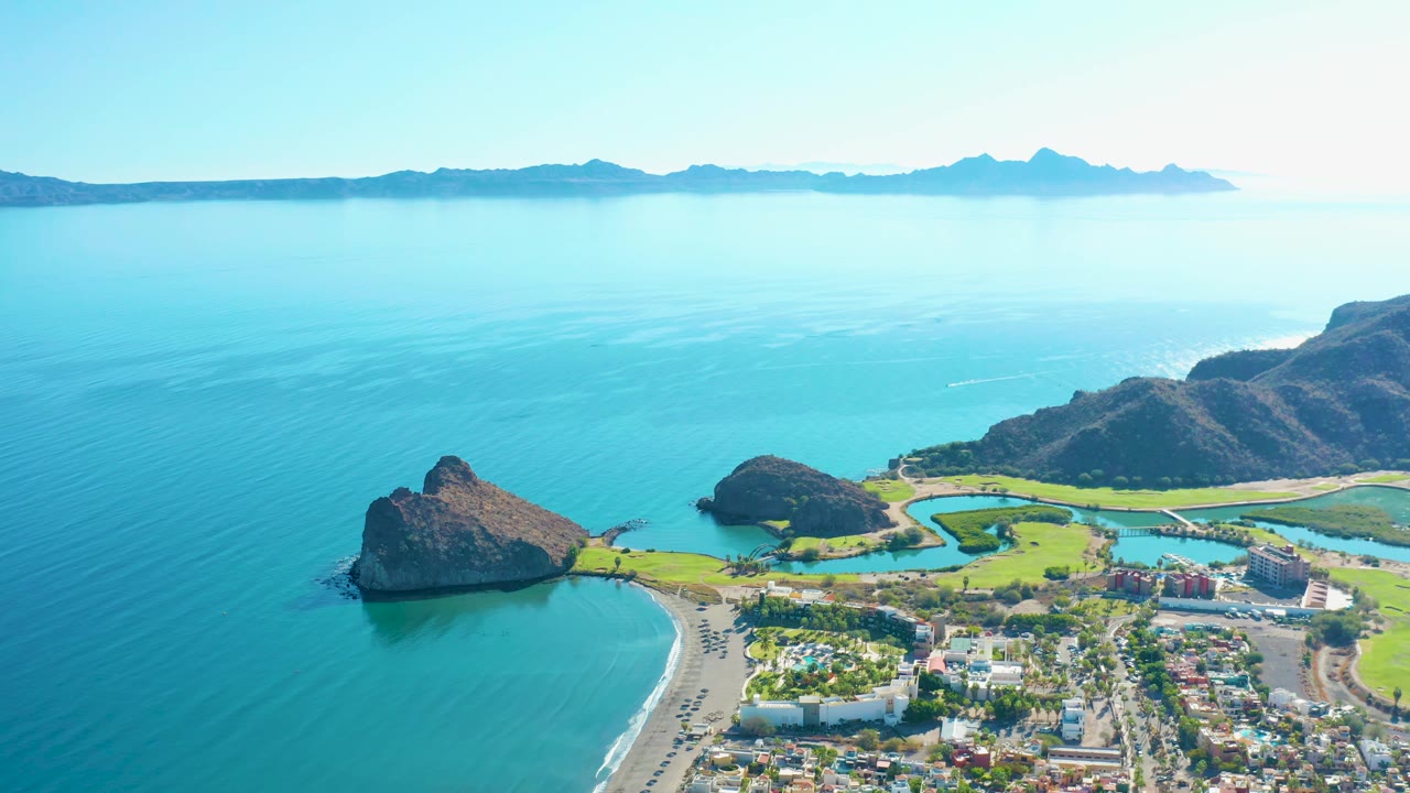 Aerial view of Loreto coastline one second into the clip, with the beach and road centered below, small vehicles on the shore road, golf-course greens near the water, hills to the right, and no visible people.
