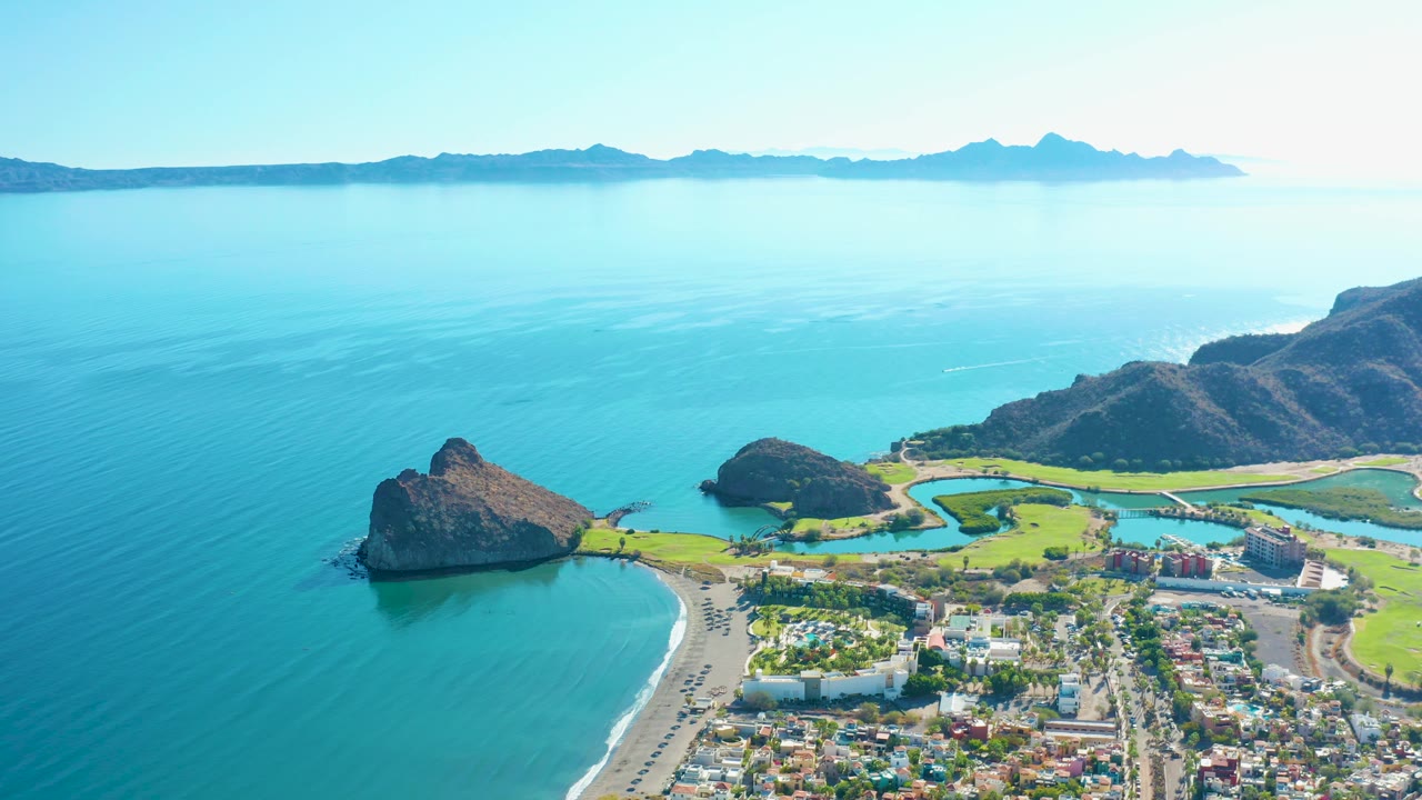 Mid-clip aerial frame over Loreto showing rocky points along the water, a pale sandy beach, the shoreline road with small traffic, fairways and buildings beside the coast, hazy distant hills, and no visible people.