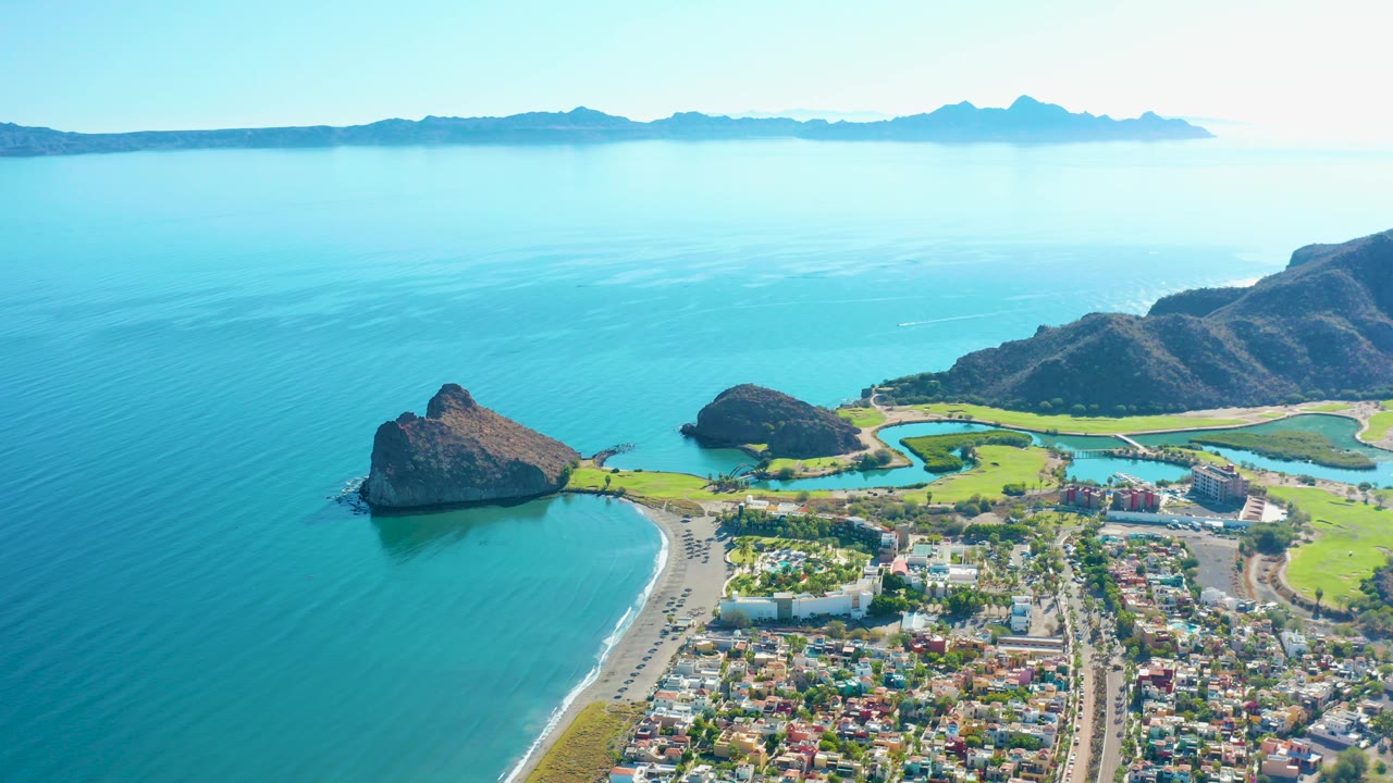 Opening aerial frame over Loreto, Baja California Sur, with turquoise bay water, a sandy beach, clustered town buildings, green fields, rocky outcrops, distant mountains, and no visible people.