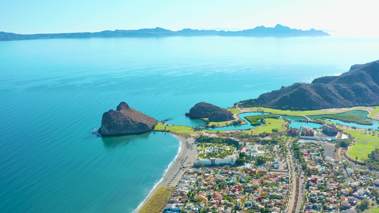 At 0:02, the Loreto aerial frames the wide turquoise bay on the left, a beach road and town below, green lagoon-like fields, rocky outcrops, a mountain ridge, and no visible people.