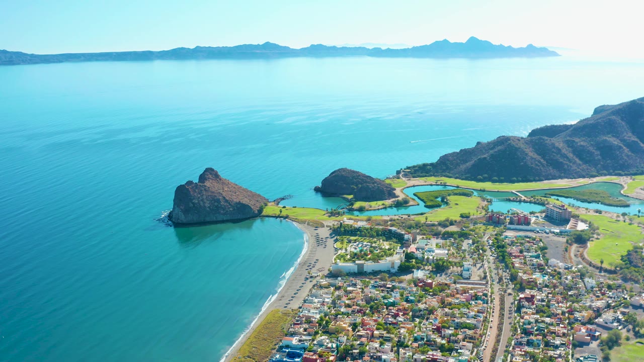 Wide aerial of calm sea, sandy shoreline, rocky point, coastal town, green fields, and distant mountains.